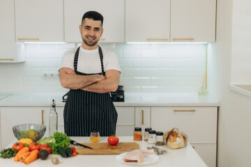 Portrait of a handsome man in the kitchen. A man dressed in a black apron. He looks at the camera. On the kitchen table are cooking products. Chicken. Vegetables. Spices