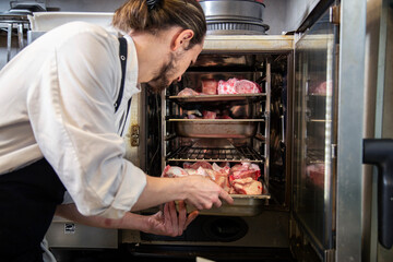 chef manos introduce bandeja con carne cruda en horno con uniforme blanco en interior de cocina de restaurante profesional detalle 