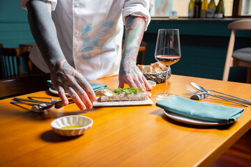 mano de chef sirve pancake de steat tartar con brotes verdes en una mesa de madera en  restaurante con copa de vino tinto y tatuajes close-up