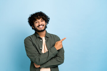 Positive indian or arabian curly haired guy in casual wear, points finger to the side at space for...