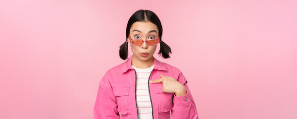 Close up portrait of asian girl looks surprised, points at herself with disbelief, being chosen, stands over pink background