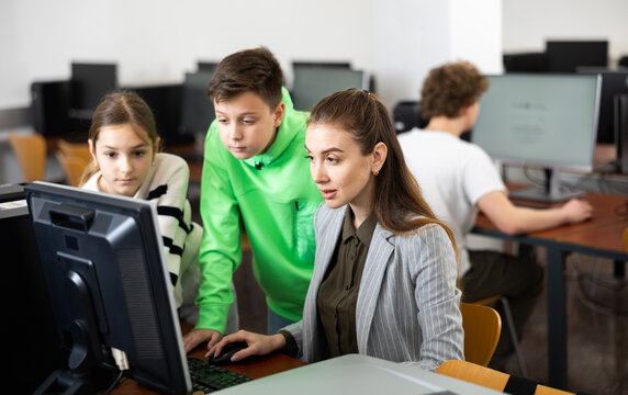 Friendly female teacher and positive teen students looking at monitor screen during lesson in computer lab at school library ..