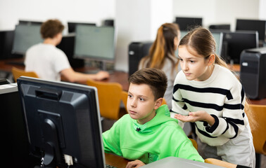 Fototapeta premium Friendly teen schoolgirl helping interested focused classmate sitting at computer in information technology class..