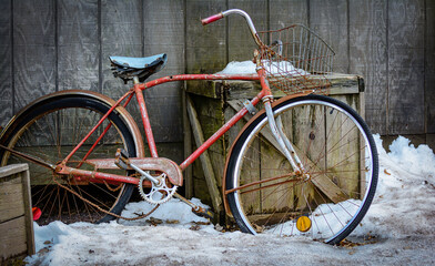 Old red bicycle abandoned in the snow on a wall of grey wood planks.