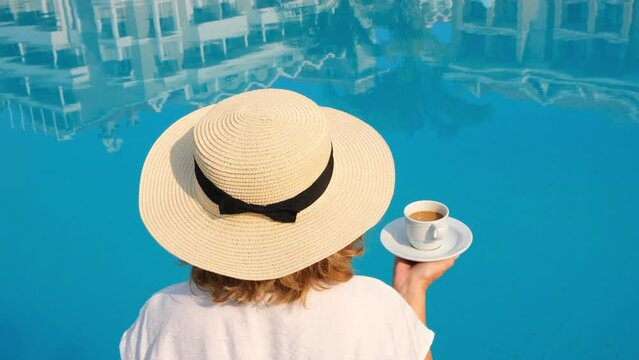 female in a straw hat holds a cup of coffee in her hand over a blue swimming pool concept of good morning and day planning, relaxation and enjoyment, back view