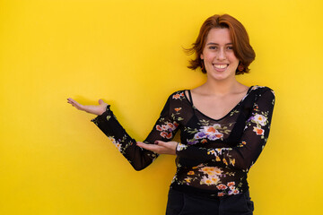 Young woman (22) Latina with positive attitude indicates with her hands a product or promotional advertisement. Space for the insertion of advertising. Yellow background. Advertising Concept