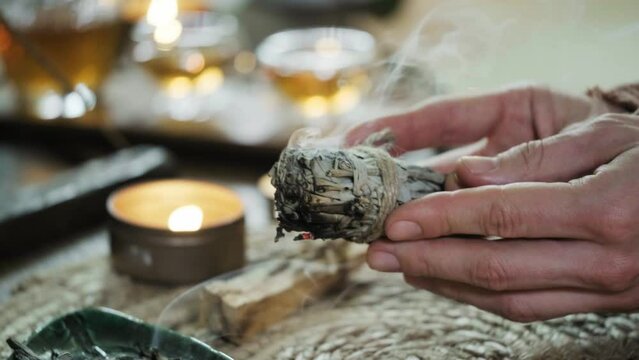 Woman hands burning white sage, before ritual on the table with candles and green plants. Smoke of smudging treats pain and stress, clear negative energy and meditation