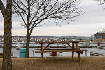 Lone picnic table at the grass at boat marina with bare tree branches. Empty boat marina  in fall. Cloudy sky.  Forest in the horizon. Gentle ripples in the water of the marina. 