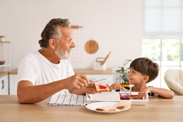Little boy and his grandfather making sandwiches with jam at table in kitchen