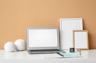 Modern laptop, smartwatch and blank photo frames on table near color wall