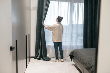 A woman with a hair bun opening the curtains in the room