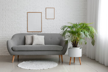 Interior of room with comfortable sofa, houseplant and blank photo frames on white brick wall