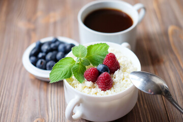 A cup of coffee and Cottage cheese in white bowl with raspberries and blueberries on wooden background.