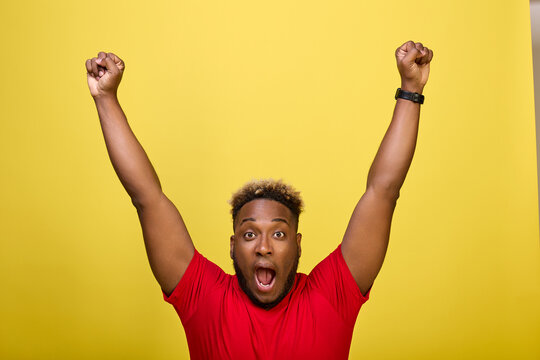Black fan rejoices at victory of his favorite team at stadium. Young black athlete in red T-shirt raised his hands happily up, holding his fists, exulting at victory of team that took first place