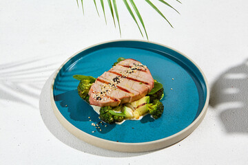 Grilled tuna steak with broccoli and zucchini in modern ceramic plate. Healthy food - roasted tuna with green vegetables. Fish dish in minimal style. Tuna fillet in blue plate with hard shadow. - Powered by Adobe