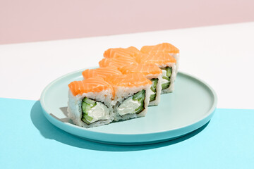 Classic sushi roll with salmon outside, cheese and cucumber inside. Philadelphia maki in minimal style. Modern japanese menu concept. Salmon maki roll on ceramic plate on coloured background.