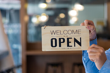 The owner holds a sign, opens the door and is ready to serve the young Asian business owner to welcome customers to the new coffee shop.