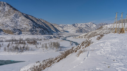 Fototapeta na wymiar A turquoise river winds through a snowy valley. Bare trees and dry grass on the banks. The poles of the power line are visible. A picturesque mountain range against the blue sky. Altai