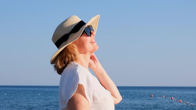 Happy mature woman 50 years old dressed in white dress, straw beach and sunglasses on the beach near the seashore. Vacation concept, relaxation, retirement age