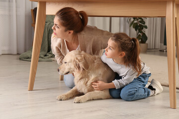 Scared mother with her little daughter and dog hiding under table in living room during earthquake - obrazy, fototapety, plakaty