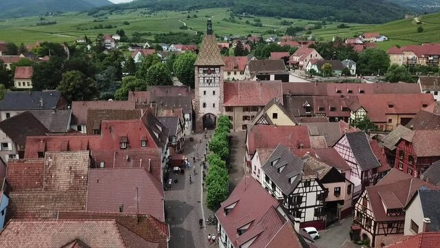 A stationary aerial footage of the clock tower surrounded by houses and the grassland. The villagers passes through the tunnel. This is located at the fortified village of Bergheim, Haut-Rhin.