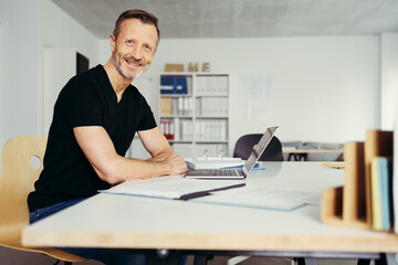 Fototapeta premium smiling man sitting in home office at desk, in front of laptop