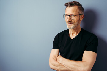 bestager with black shirt and glasses stands in front of a blue wall and looks to the side, arms...
