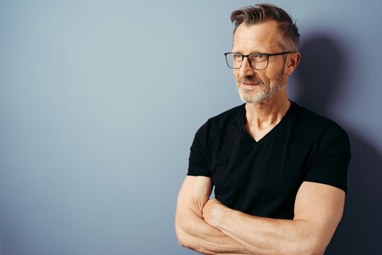 bestager with black shirt and glasses stands in front of a blue wall and looks to the side, arms crossed