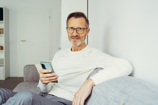 older man sits on sofa with cell phone and glasses and looks into camera