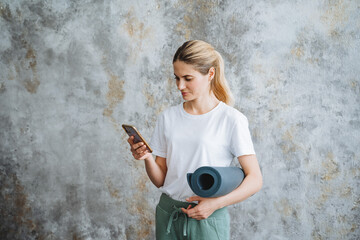 Phone in hand, young beautiful girl typing text on a smartphone, holding a yoga mat in her hands,...