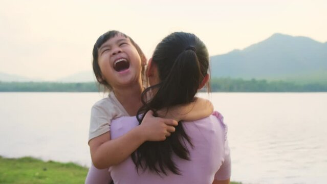 Mother holding a cute little daughter walking by the lake at sunset. Excited kid girl laughs playing with her mother outdoors. Mother hugging her daughter.