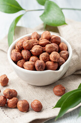 Peeled hazelnuts in a white cup. Gray linen background. Green leaves in the background.