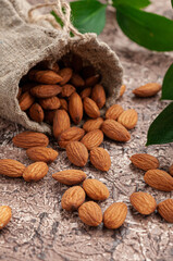 Almonds are scattered on the table. In the background is a bag of almonds and green leaves. Brown background. Close-up.