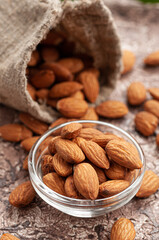 Almonds in a glass bowl. In the background is a bag of almonds and green leaves. Brown background. Close-up.