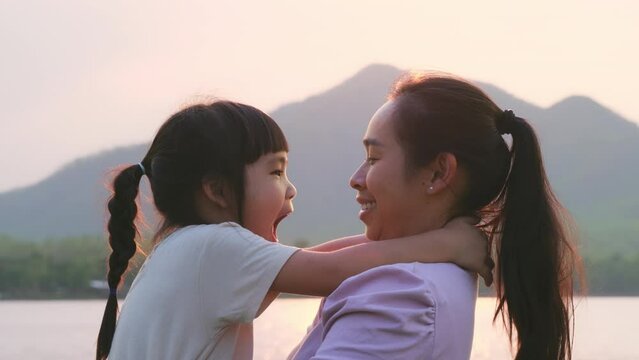 Mother holding a cute little daughter walking by the lake at sunset. Excited kid girl laughs playing with her mother outdoors. Mother hugging her daughter.