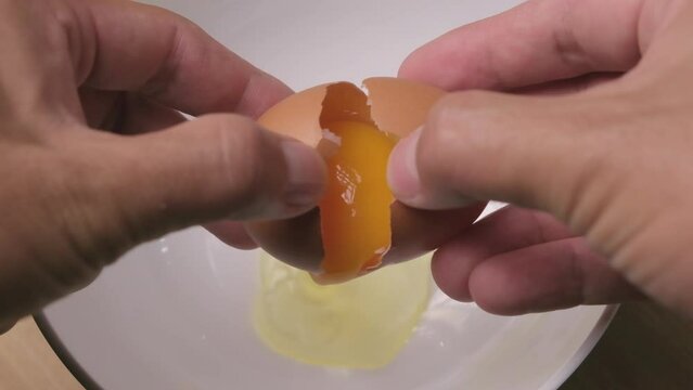 Slow motion of a hand cracking an egg into a white bowl reveals the yolk. Cooking breakfast. Step-by-step process of making omelettes. Healthy food concept.