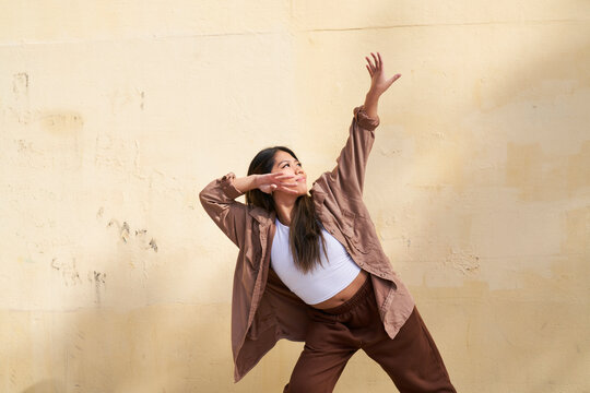 Woman dancing in front of beige wall with hand to cheek and arm raised