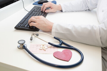 Doctor enters information about the patient and the results of the examination. There is a stethoscope, a cardiogram of the heart and pills on the table. Red heart. Examination by a cardiologist.