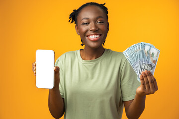 Young black woman holding white screen smartphone and money in yellow studio