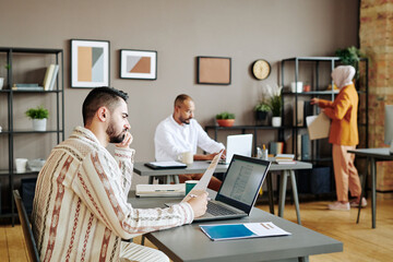 Obraz premium Serious young male economist analyzing financial papers in front of laptop by workplace in office against two busy coworkers