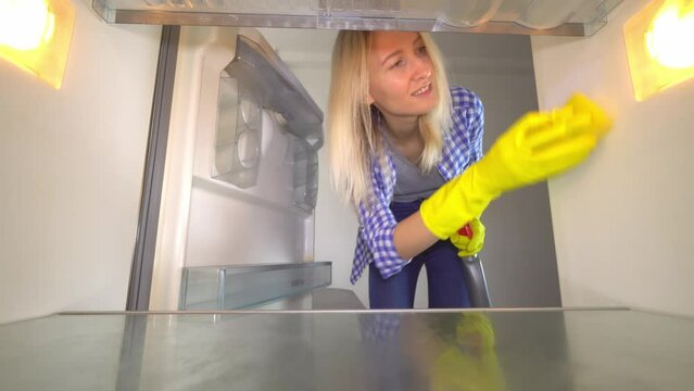 An inside view of a woman in rubber gloves cleaning an empty refrigerator. Housewife, slender blonde Mila White, washes refrigerator with detergent spray and sponge. Wide viewing angle. Smile and joy.