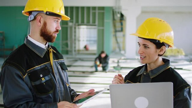 In front of the camera young man architect and the woman engineer in safety helmets and special uniforms discussing in the inside of construction building. 4k