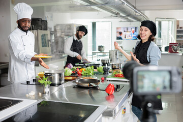 Fototapeta na wymiar Food industry workers shooting video for television cooking show while creating fine dining gourmet dish. Chefs in restaurant kitchen recording preparation process of meal for internet course.