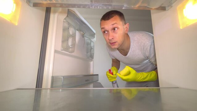young white European opens refrigerator to wash it. Wearing gloves, with a sponge and spray from sniffing and plugging the nose with your hand because of the terrible smell. Germs and freon leakage.