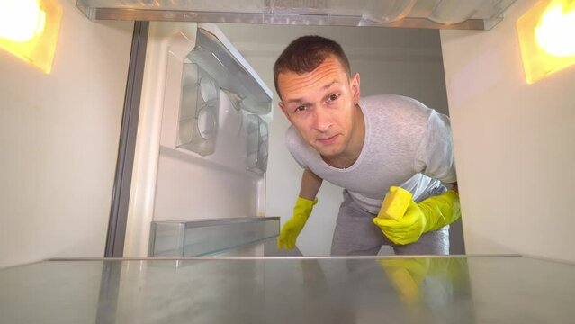 young white European opens refrigerator to wash it. Wearing gloves, with a sponge and spray from sniffing and plugging the nose with your hand because of the terrible smell. Germs and freon leakage.