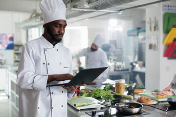 Gourmet cuisine head chef with laptop looking for gastronomic dish recipes and garnish ideas in restaurant kitchen. Gastronomy expert wearing professional cooking uniform while searching meal ideas.