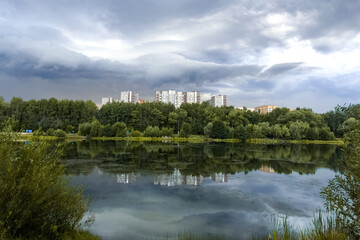 Summer city landscape with calm lake water surface with cloudy sky and multi- storey residential buildings reflected on water