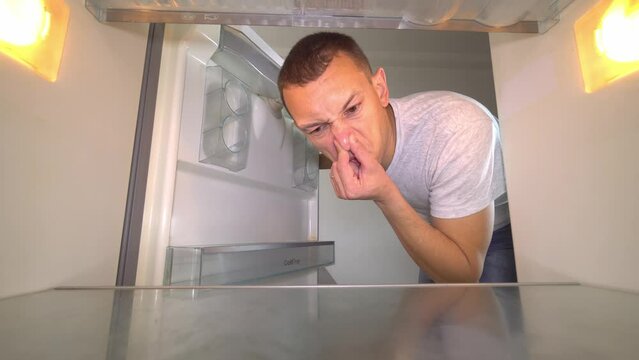 young white European opens refrigerator to wash it. Wearing gloves, with a sponge and spray from sniffing and plugging the nose with your hand because of the terrible smell. Germs and freon leakage.