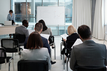 Group of contemporary economists analyzing financial documents with data while sitting in rows in front of whiteboard in auditorium