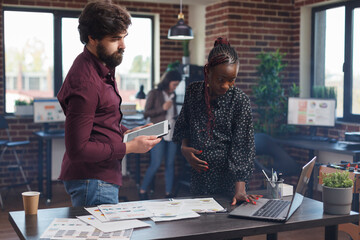 Multi ethnic diverse company business employees trying to solve financial issues using tablet and laptop. Expectant mother working on modern computer while analyzing marketing strategy ideas.